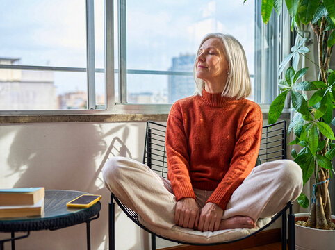 Content senior woman with closed eyes enjoying warm sunlight indoors, meditating or relaxing in peaceful home.