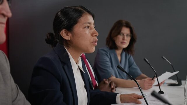 Young Hispanic woman speaking into microphone during international summit panel with multiethnic colleagues. Footage is perfect for political news reports side view shot