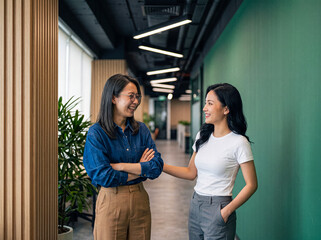 Naklejka premium Two happy young Asian female colleagues smiling, sharing a friendly conversation and positive interaction in a bright.