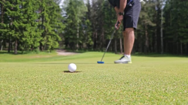 Golf player putts golf ball towards hole on course under sunlight near trees in afternoon