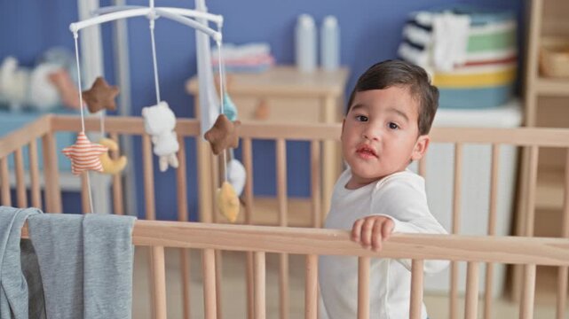 Little boy stands in white onesie, grips crib rail and reaches hand to hanging mobile in nursery building; gentle curiosity.