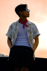 Fototapeta premium Stylish young Asian man in retro summer outfit posing outdoors against golden sunset sky background