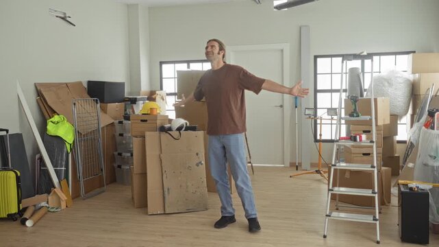 Man arms outstretched amid stacked cardboard boxes, ladder, tools and bubblewrap in a studio; fresh start relief.