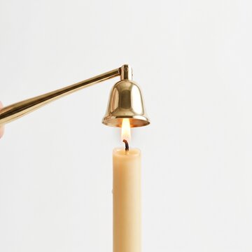 Close up of a hand holding a brass candle snuffer over a lit beeswax candle against a white background