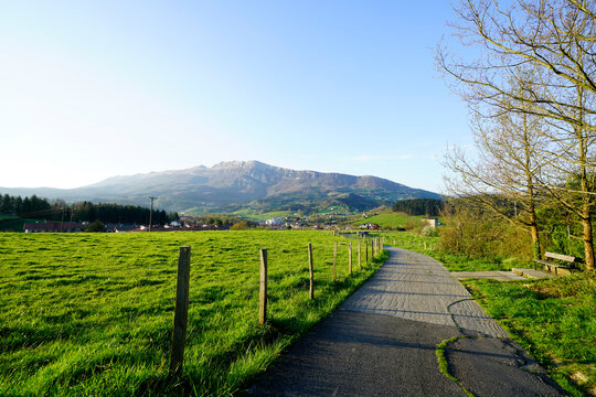Nature landscape of green grass field, walkway, mountain, and blue sky on sunny day in spring. Cow farming ranch. Animal pasture. Asphalt path beside fence of farmland. Green environment in Europe.
