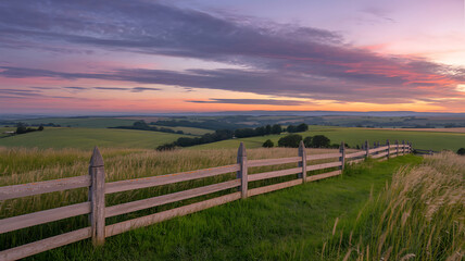 Obraz premium A wooden fence in a green field at sunset, rolling hills and trees in the background