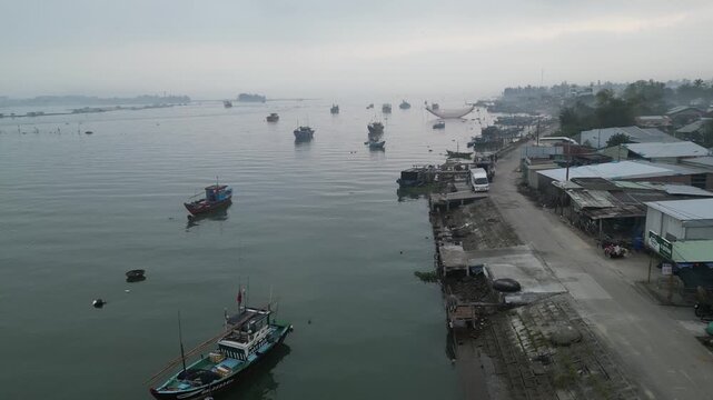 Aerial view of traditional fishing boats and round basket boats moored along the misty riverbank with local buildings and a coastal road in Hoi An, Da Nang, Vietnam.