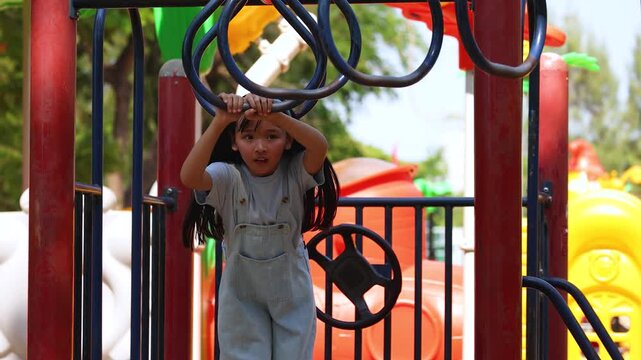 Young Thai Girl in Blue Overalls Climbing Monkey Bars at Playground
