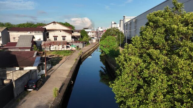 Drone view of urban drainage canal with dark water reflection, concrete and wooden retaining walls beside narrow road and old buildings. Urban waterway, city infrastructure and flood control concept.