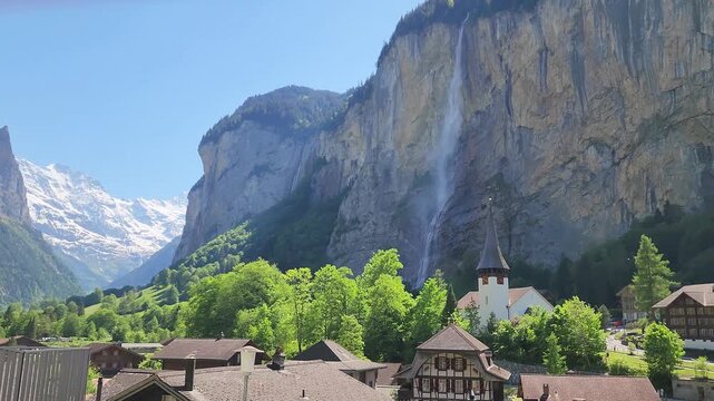 Lauterbrunnen, Switzerland, panning camera movement of an idyllic alpine village with waterfall, bell tower and snow covered mountains on a summer day.