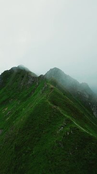 Aerial view of a narrow hiking trail winding along a steep green mountain ridge with mist rolling over the peaks and a hiker walking in Martigny, Valais, Switzerland.