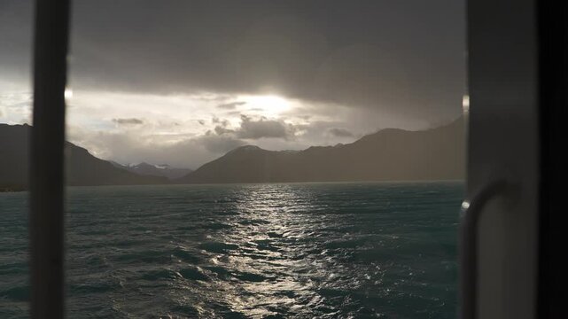Lago Argentino, Los Glaciares National Park, Santa Cruz, Argentina, featuring rough turquoise waters, mountain silhouettes, rain shafts and reflected sunlight beneath storm clouds, slow motion lateral