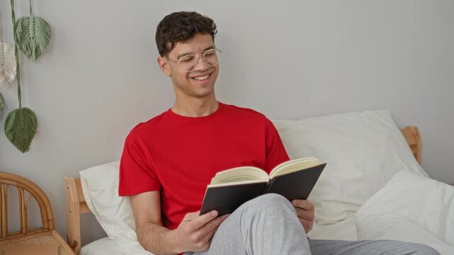 Man in red shirt reading book on bed, holding open book and smiling inside a building bedroom; calm focus.