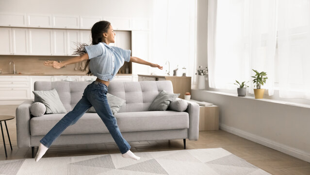 Beautiful preteen age girl having fun in cozy living room, spread her arms and legs while jumping, looks like ballerina, rehearsing, performing dance enjoy active carefree leisure alone at modern home