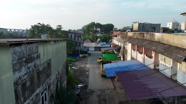 A narrow urban alley, lined with corrugated-metal warehouses, parked delivery trucks, and makeshift awnings, paints a picture of a rugged industrial area with dilapidated buildings.