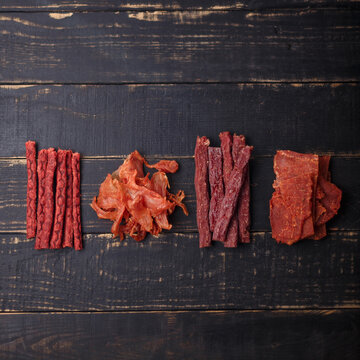 Dry salmon fish, chicken, red beef sticks and pork slices. Jerky meat on black wooden background. No brand, product presentation. Copy space, top view.