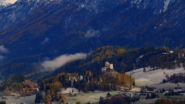 Aerial view of the historic Schloss Tarasp perched on a rocky hill surrounded by autumn forests and snow-capped mountains in Scuol, Graubunden, Switzerland.