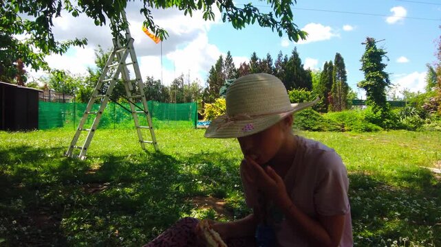 Outdoor medium static shot captures a young girl wearing a straw hat sitting on the grass and eating a snack during a lunch break in a sunny backyard orchard garden with a metal ladder.