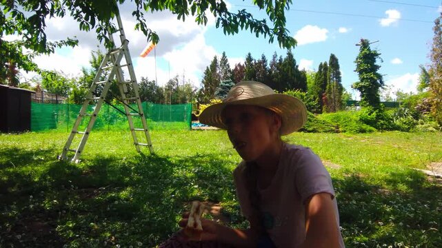 Outdoor wide static shot captures a tired young girl wearing a straw hat sitting on the grass and eating a snack during a lunch break in a sunny backyard orchard garden with a metal ladder.