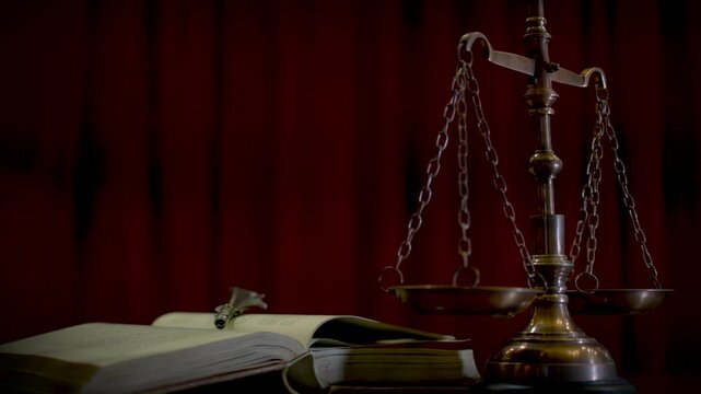 A side view of a brass scale of justice on a wooden surface with a deep red curtain in the background and warm dramatic lighting.