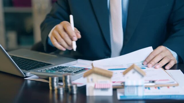Businessman holding a pen, pointing at a house model and financial charts to review property contract details.