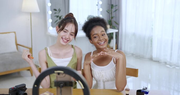 asian teenage beauty content creator with african american teenage girl laughing sharing makeup palette during slowmotion video tutorial shoot at bright home studio with skincare bottles camera setup