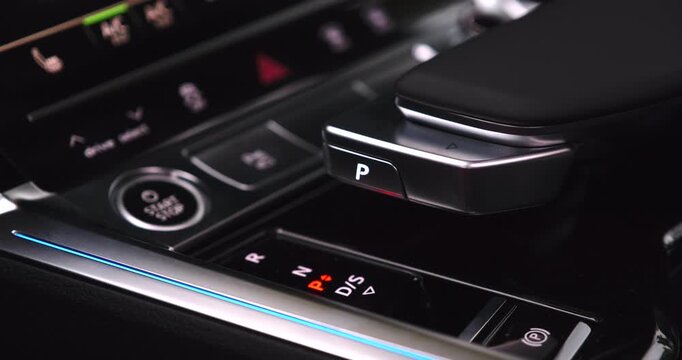 Close-up of a driver pushing the park button on a modern automatic transmission gear selector. The elegant interior features ambient lighting
