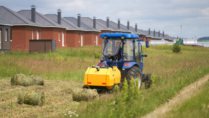 Obraz premium A compact tractor with a miniature hay baler attached works on a narrow strip of field on the outskirts of a village.