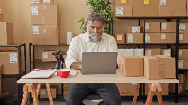 Man middle aged with beard scans parcel with barcode scanner while holding box beside laptop at packing desk in building; efficiency focus.