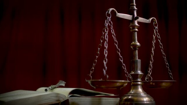 A brass scale of justice on a wooden desk with a dark red curtain background shot from a low angle with dramatic warm lighting.