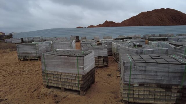 The area shows many pallets stacked with building materials arranged neatly in rows. The ground is sandy, and there are piles of earth nearby. Dark clouds fill the sky, indicating the possibility of r