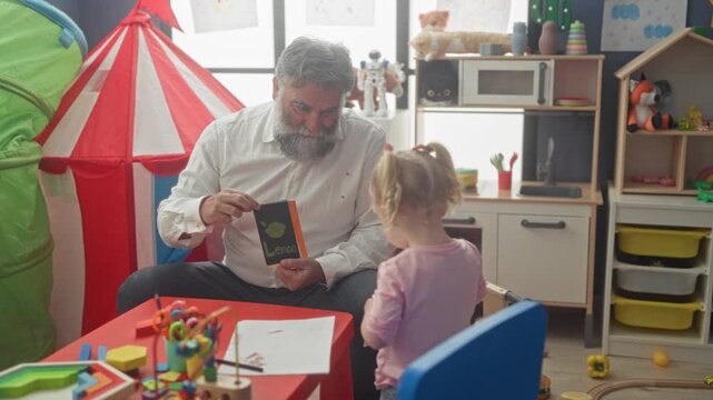 Man shows illustrated flashcards of cat and dog to little girl in bright playroom, seated at a red table with toys and tent; nurturing learning bond.