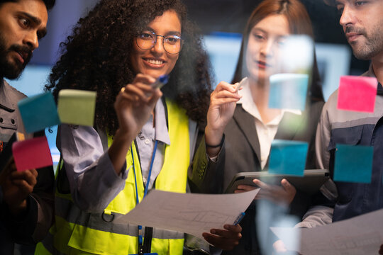 Smiling Black female engineer writes ideas on a sticky-note glass board while three diverse team members observe in a dark collaborative workspace.