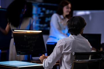 Red-haired female professional in a cream jacket holds documents and speaks to a Black female analyst beside a glowing monitor in a dark tech room. © ultramansk