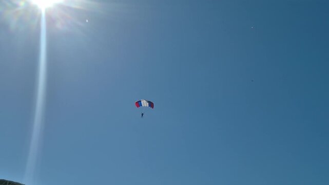 Skydiver in parachute descending against clear blue sky. Professional paraglider falling to landing. Extreme sport lifestyle activity, recreational air descent.