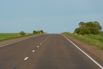 Fototapeta premium Long straight asphalt road leading toward the horizon through open fields under clear sky