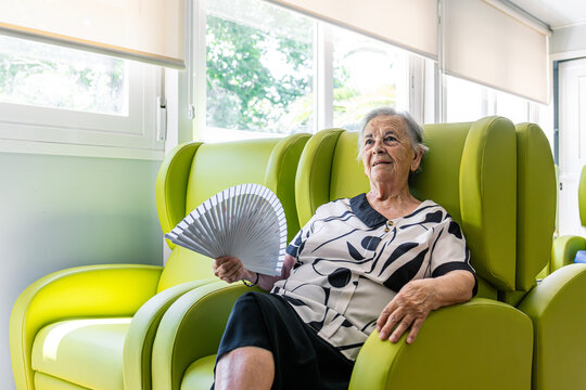 Senior woman cooling off with a hand fan while relaxing in a lounge chair at a retirement home, serene and comfortable indoors