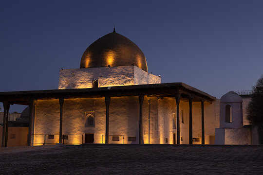 Historic domed building illuminated at twilight inside Ichan Kala old town in Khiva, Uzbekistan. Warm uplighting highlights whitewashed walls and wooden columns against a deep blue evening sky.