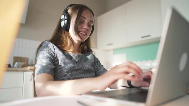 Woman typing on laptop wearing headphone at home. Freelancer works remotely on computer in kitchen. Woman with headphone uses laptop at desk. Remote worker types on notebook. Smiling woman at home.