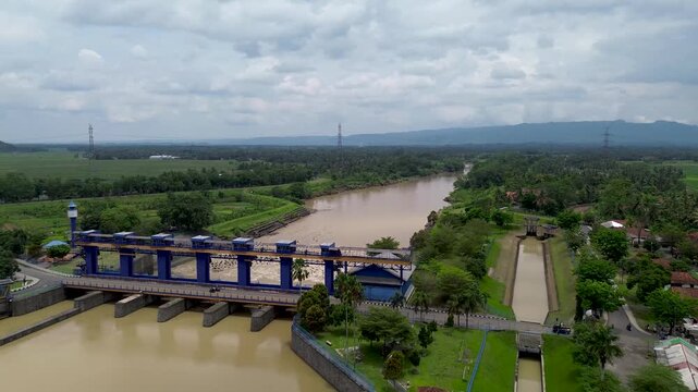 An aerial perspective showing the large water control gates of the Manganti Barrage, surrounded by lush green landscapes and the Serayu River in Indonesia.