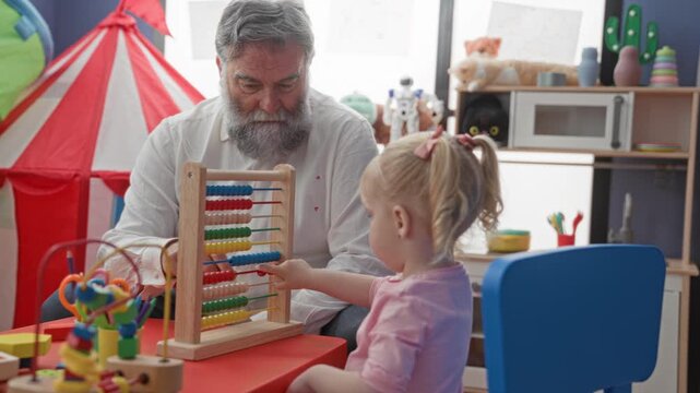 Man watches little girl point finger to beads on a wooden abacus at a colorful preschool building play table; nurturing learning bonding.