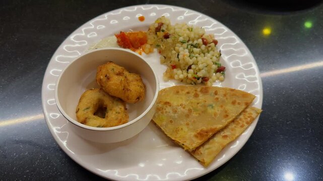 Camera orbiting around a plated South Indian breakfast featuring golden paratha wedges, crispy medu vada, and sabudana khichdi served on a white plate under indoor lighting.