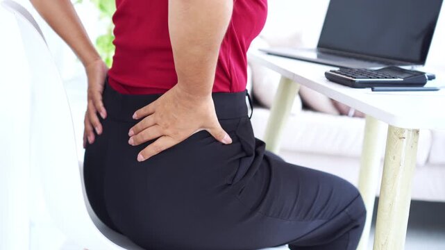 close-up of a woman sitting at her office desk holding her painful lower back, suffering from backache and office syndrome while working with a laptop and a calculator on a white tabl