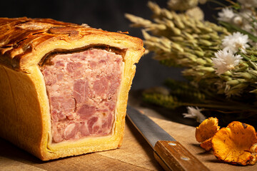 Obraz premium Pate en croute or pâté en croûte on a wooden cutting board, aside a knife and Chanterelle mushrooms on a dark gray background with dried flowers. Countryside vintage atmosphere.
