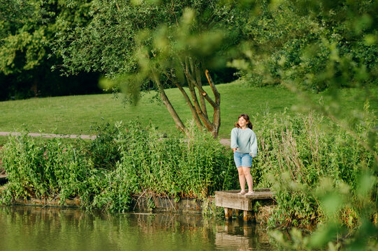 School age girl standing alone on small dock beside green lakeshore