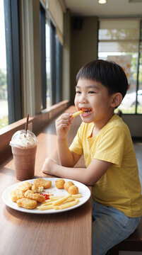 Side view of cute Asian boy smiling while eating french fries and chicken nuggets with chocolate smoothie covered with whipped cream on wooden table at cafe