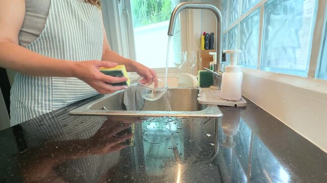 Woman washing a glass salad bowl in a kitchen sink with running water, everyday housework and home cleaning routine.