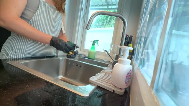 Woman cleaning and rinsing a kitchen sink drain strainer with a sponge, everyday household cleaning and hygiene routine in a modern kitchen.