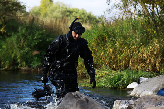 Underwater Videographer in Black Wetsuit Navigating River Rocks Amidst Green Foliage