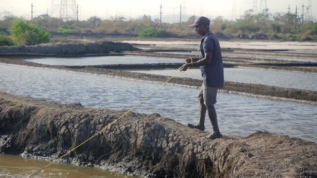 Daylight outdoor shot of an Indian Asian male fisherman casting a fishing net in the pond, lake, or canal water in search of fish on a sunny but windy day with salt pans visible in the background. 